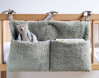 May include: A light green, fuzzy, hanging organizer with two pockets. The organizer is attached to a wooden crib with grey fabric ties. A stuffed animal is in one pocket and a book titled "Clap Hands" is in the other pocket.