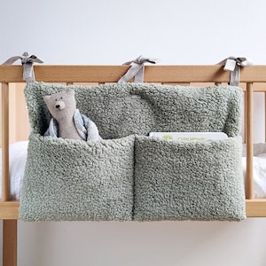 May include: A light green, fluffy hanging organiser with two pockets. The organiser is attached to a wooden cot with grey fabric ties. A stuffed toy is in one pocket and a book titled "Clap Hands" is in the other pocket.