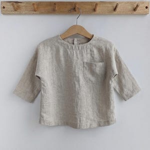 May include: A beige, long-sleeved shirt with a single pocket. The shirt is made of a textured fabric and is hanging on a wooden hanger. The shirt is displayed against a white background. The shirt's sleeves are three-quarter length.