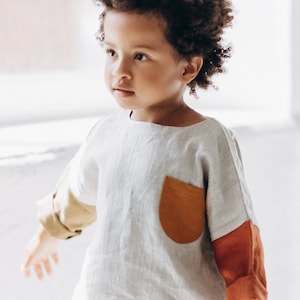 Peut inclure: Un jeune enfant portant une chemise en lin beige clair avec une grande poche orange et des manches en patchwork coloré. L'enfant a les cheveux courts et bouclés bruns et regarde vers la gauche.