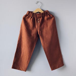 May include: Rust-colored linen pants with an elastic waistband and two button closure. The pants are hanging on a wooden hanger against a gray background. The pants have a relaxed fit and a simple design.