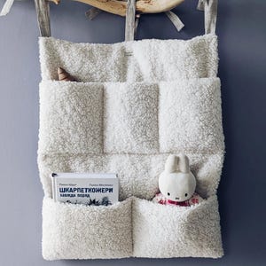 Op de afbeelding: Witte, pluche wandorganizer van stof met zes vakken. De organizer hangt aan een tak. Een boek met de titel "ШКАРПЕТКОЖЕРИ" zit in een vak en een wit knuffelkonijn in een ander vak.