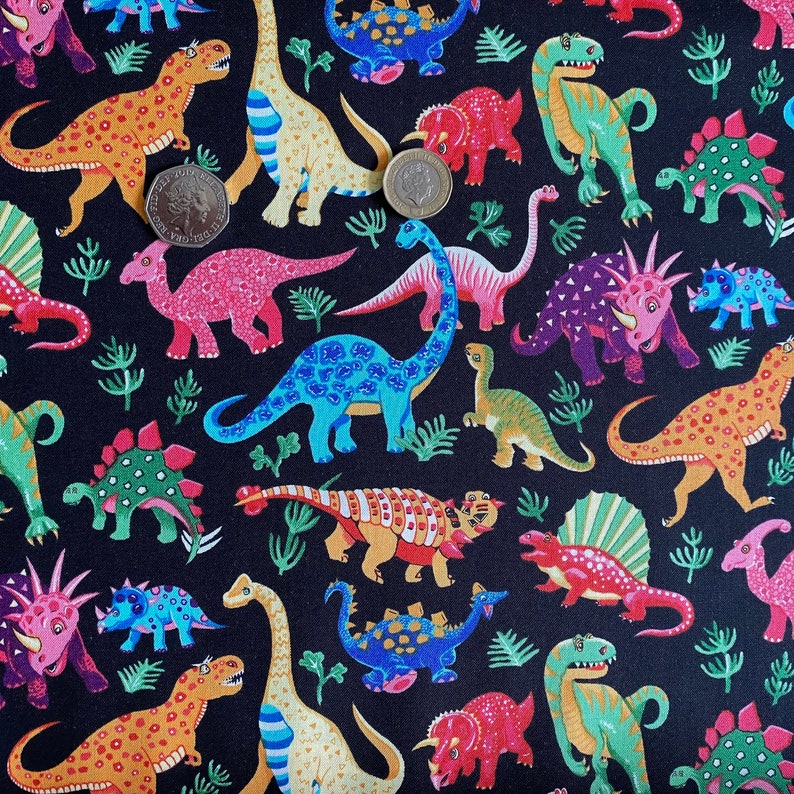 May include: A black fabric featuring a colorful dinosaur pattern. The dinosaurs are in various colors, including orange, blue, green, and pink, with a few sprigs of green foliage scattered throughout. The dinosaurs are of different species and sizes.