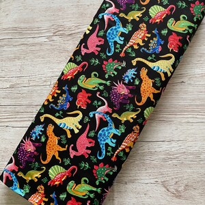 May include: A black fabric featuring a colorful dinosaur pattern. The dinosaurs are in various colors, including pink, blue, yellow, orange, and green. The fabric is likely cotton or a similar material, suitable for quilting or crafting projects.