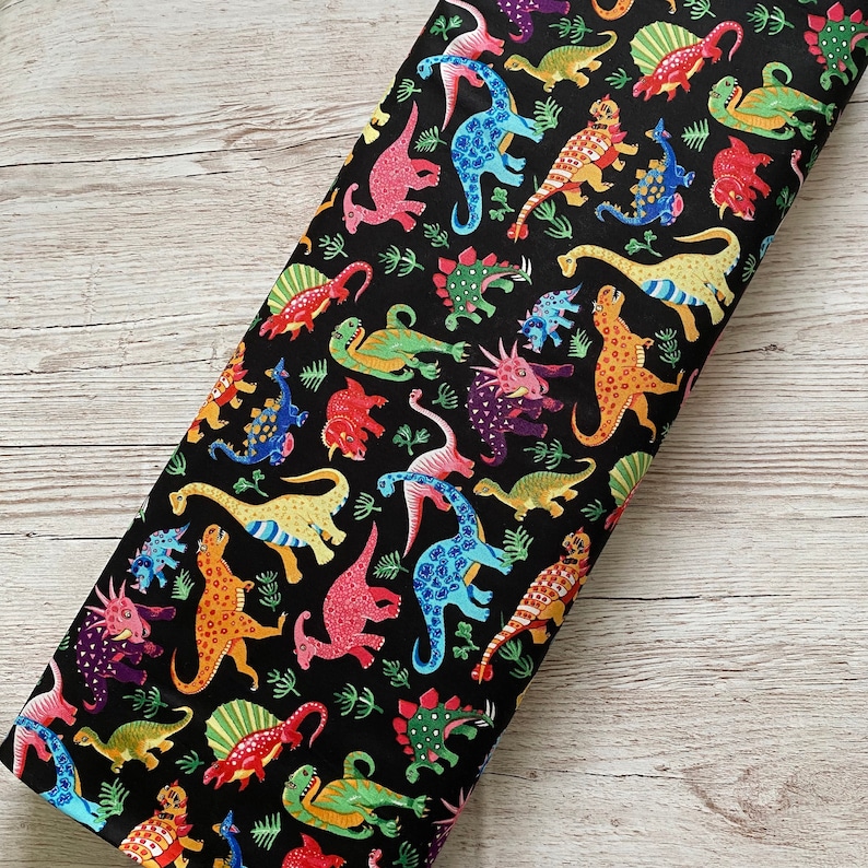 May include: A black fabric featuring a colorful dinosaur pattern. The dinosaurs are in various colors, including pink, blue, yellow, orange, and green. The fabric is likely cotton or a similar material, suitable for quilting or crafting projects.
