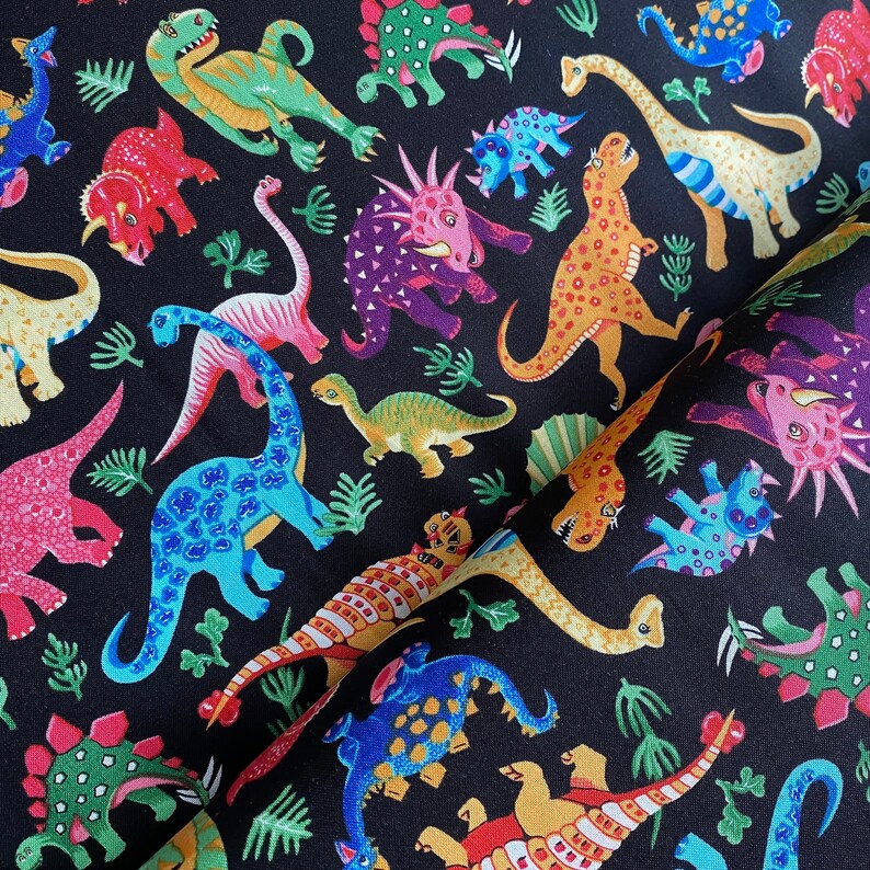 May include: Black fabric featuring a colorful dinosaur pattern. The dinosaurs are depicted in various colors, including red, green, blue, and yellow, with a variety of shapes and sizes. The fabric also includes green plant illustrations.