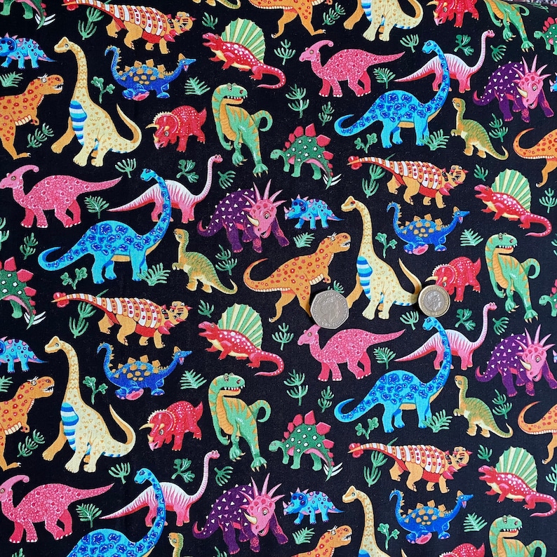 May include: Black fabric with a colorful dinosaur pattern. The dinosaurs are in various colors, including orange, blue, pink, and green, with some having patterned designs. The background is black, and there are small green plant-like shapes scattered throughout.