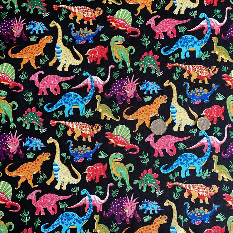May include: A black fabric covered with colorful cartoon dinosaurs in various poses. The dinosaurs are in shades of red, orange, blue, green, and purple. Small green plant-like shapes are scattered throughout the design.