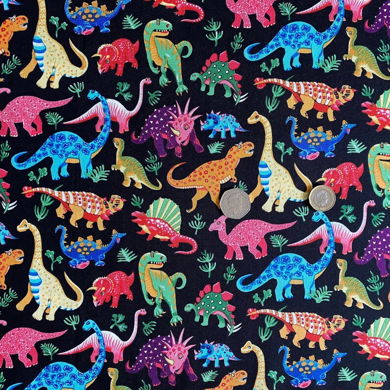 May include: A black fabric featuring a colorful dinosaur pattern. The dinosaurs are in various colors, including blue, pink, green, and orange, with some having spots and stripes. The design includes ferns and various dinosaur species.