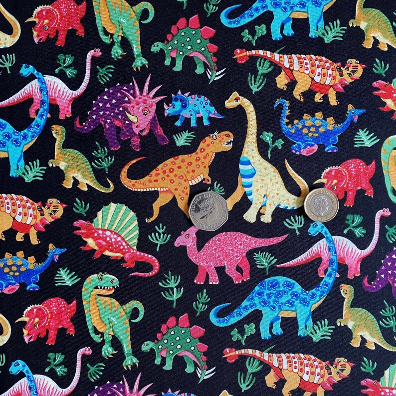 May include: Black fabric featuring a colorful dinosaur pattern. The dinosaurs are in various colors, including red, green, blue, and orange, with a few small green plant illustrations. The fabric has two coins on it.