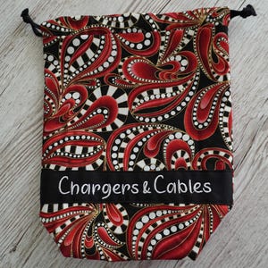May include: A drawstring bag with a black background and a red and white paisley pattern. The bag has a black band with the words "Chargers & Cables" embroidered in white. The bag is designed to hold chargers and cables.