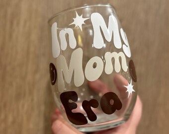 May include: A clear glass wine glass with the text "In My Mom Era" in white and brown lettering with stars.