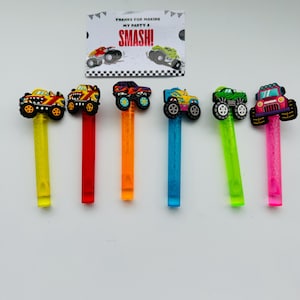 Op de afbeelding: Zes kleurrijke monster truck party favors met bijpassende gekleurde stelen. Elke truck heeft een andere kleur en een ander ontwerp. Een kleine kaart op de achtergrond leest "Bedankt voor het maken van mijn feest een succes!"