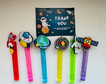 Può includere: Sei bacchette di plastica colorate con diversi topper a tema spaziale, tra cui un razzo, un'astronave, un pianeta, un sistema solare, un astronauta e un satellite. Le bacchette sono su una superficie bianca con un biglietto di ringraziamento per una festa di compleanno.