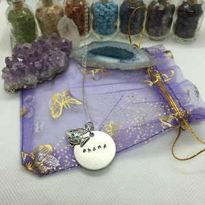 May include: A collection of crystals, gemstones, and a necklace with the word "ohana" stamped on a silver pendant. The necklace features a turtle charm with blue and green accents. Small glass bottles filled with colorful stones are arranged in the background.
