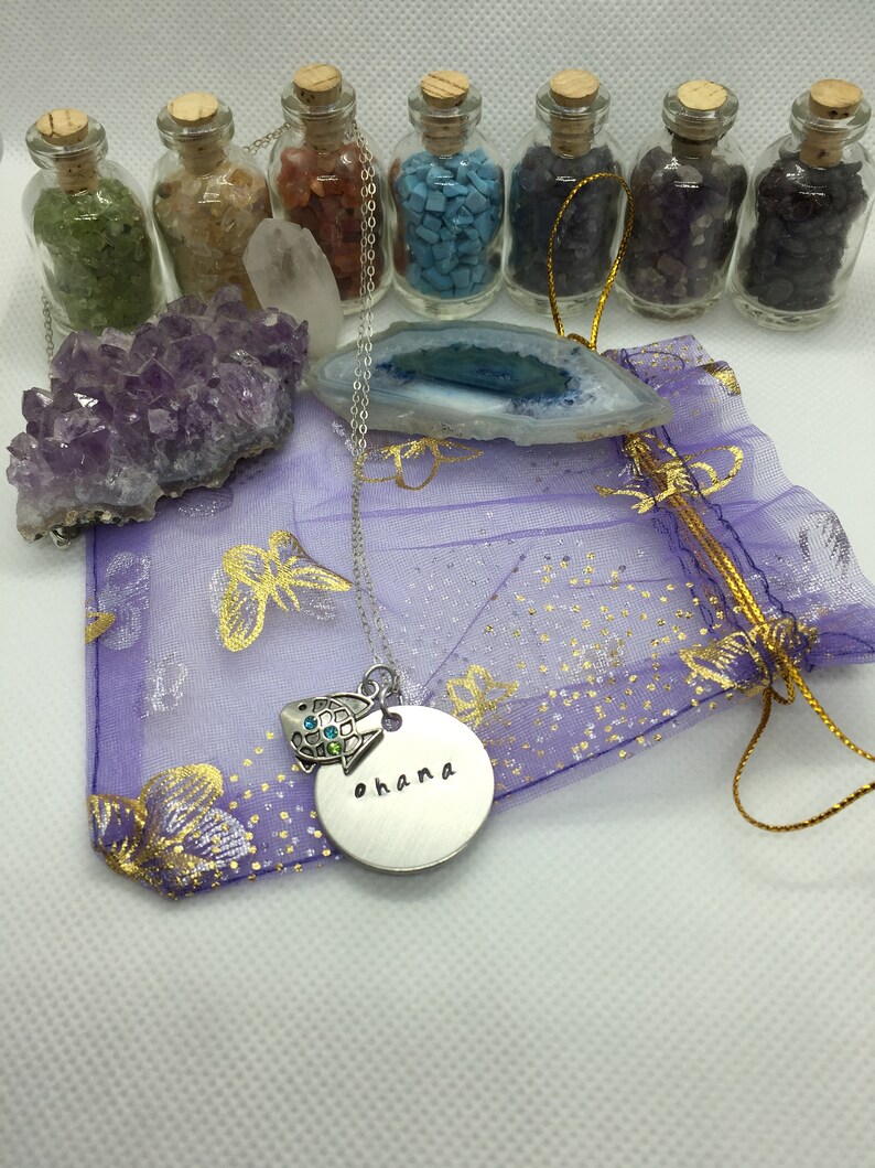 May include: A collection of crystals, gemstones, and a necklace with the word "ohana" stamped on a silver pendant. The necklace features a turtle charm with blue and green accents. Small glass bottles filled with colorful stones are arranged in the background.