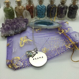 May include: A silver necklace with a turtle charm and a round pendant that says "ohana" in black lettering. The necklace is on a purple bag with gold accents, with small glass bottles filled with colorful stones in the background.