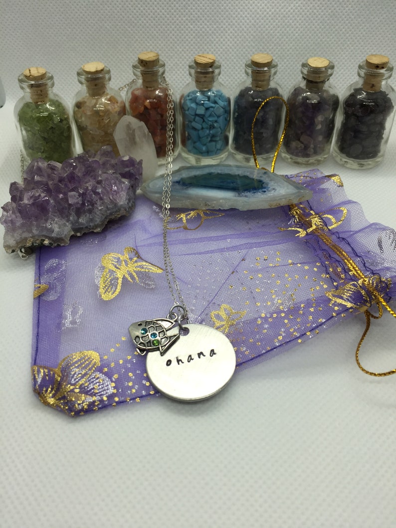 May include: A silver necklace with a turtle charm and a round pendant that says "ohana" in black lettering. The necklace is on a purple bag with gold accents, with small glass bottles filled with colorful stones in the background.