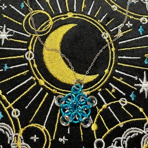 Op de afbeelding: Een zilveren ketting met een blauw-zilveren bloemhanger. De hanger is gemaakt van in elkaar grijpende metalen ringen. De ketting wordt gepresenteerd op een zwarte achtergrond met een gouden halve maan en sterrenontwerp.
