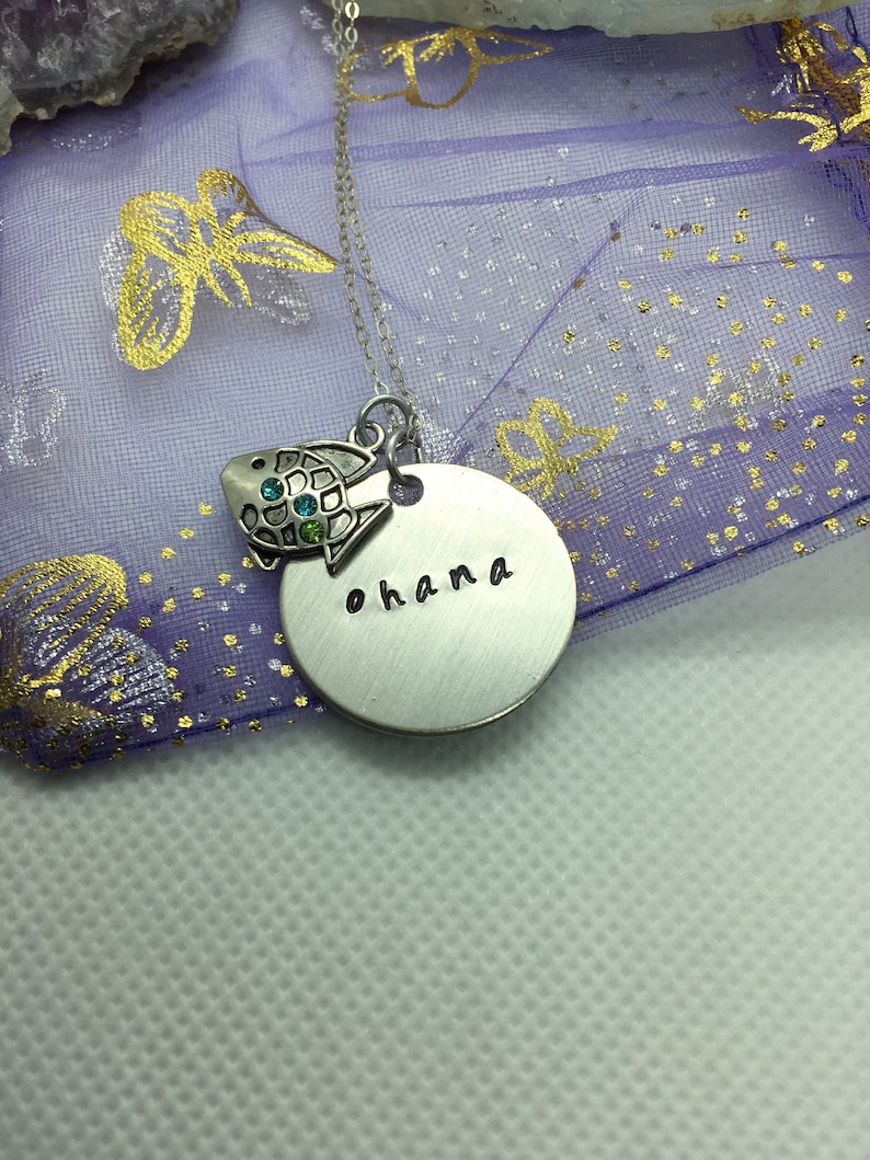 May include: A silver-toned necklace featuring a round pendant stamped with the word "ohana." A small, decorative turtle charm with blue and green accents hangs next to the pendant. The necklace is displayed on a purple fabric with gold accents.