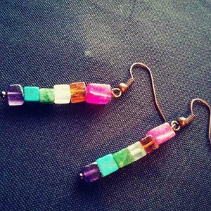 Puede incluir: Un par de pendientes con un diseño de piedras de colores. Cada pendiente presenta una serie de pequeñas piedras cuadradas en tonos de morado, turquesa, verde, blanco, naranja y rosa.