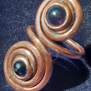 May include: A copper ring with two spiral designs, each featuring a dark purple gemstone in the center.