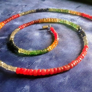 Könnte beinhalten: Eine mehrfarbige Perlenkette mit abwechselnden Farben in Rot, Orange, Gelb, Grün, Blau und Schwarz. Die Kette besteht aus kleinen, runden Perlen und ist auf einer silbernen Kette aufgefädelt.