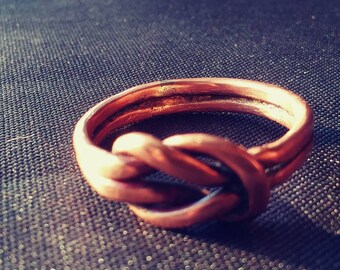 Puede incluir: Un anillo de cobre con un diseño de nudo. El anillo está hecho de un solo alambre que se tuerce y anuda.