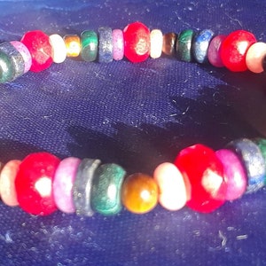 Puede incluir: Un brazalete de cuentas de colores con cuentas rojas, blancas, verdes, azules y doradas. El brazalete está hecho de varios materiales, incluyendo cuentas de piedras preciosas y cuentas de metal.
