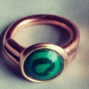 Könnte beinhalten: Ein Kupferring mit einem grünen Stein, der in einer goldenen Fassung sitzt. Der Stein hat ein dunkelgrünes Design in der Mitte.