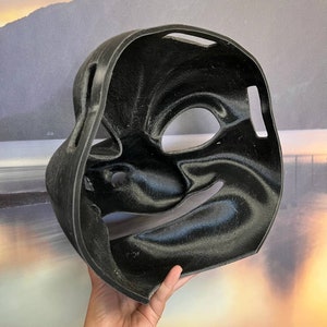 Puede incluir: M&aacute;scara de pl&aacute;stico negro con una superficie texturizada y aberturas para los ojos y la boca. La m&aacute;scara est&aacute; dise&ntilde;ada para parecerse a un rostro humano.