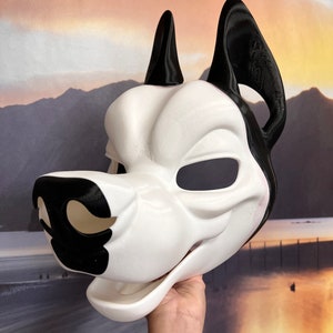 May include: A white and black dog mask with large, pointed ears. The mask has a smooth, matte finish and features black accents on the nose and ears. The eye openings are dark, and the mouth is open. The mask is held up by a hand.