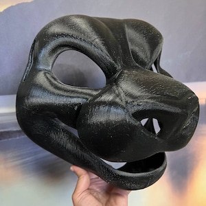 Puede incluir: Una m&aacute;scara de animal negra impresa en 3D con un hocico detallado y la boca abierta. La m&aacute;scara est&aacute; hecha de un material liso y texturizado.