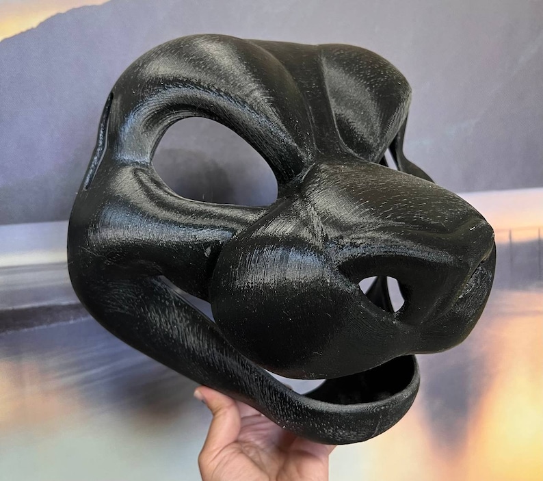 Puede incluir: Una m&aacute;scara de animal negra impresa en 3D con un hocico detallado y la boca abierta. La m&aacute;scara est&aacute; hecha de un material liso y texturizado.