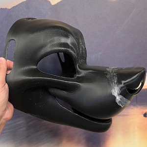 Op de afbeelding: Zwart bont dierlijk masker met een gedetailleerde snuit en open mond. Het masker is gemaakt van een glad, hard materiaal en heeft een getextureerd oppervlak.