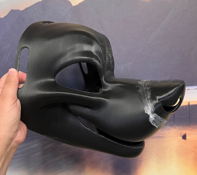 Op de afbeelding: Zwart bont dierlijk masker met een gedetailleerde snuit en open mond. Het masker is gemaakt van een glad, hard materiaal en heeft een getextureerd oppervlak.