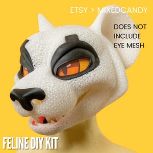 May include: A white feline mask base for a DIY kit featuring black eye markings, a black nose, and orange eyes. The text on the image reads: ETSY > MIXEDCANDY, DOES NOT INCLUDE EYE MESH, and FELINE DIY KIT. The mask has a textured surface.