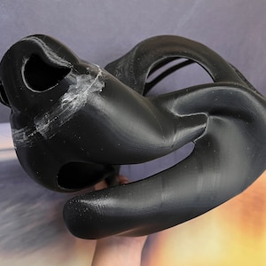 Op de afbeelding: Zwart 3D-geprint masker met een grote, open mond en een kleiner, open ooggat. Het masker is gemaakt van een glad, plastic materiaal.