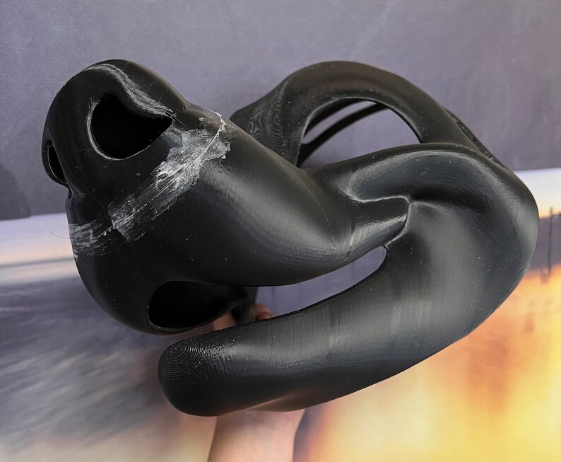 Op de afbeelding: Zwart 3D-geprint masker met een grote, open mond en een kleiner, open ooggat. Het masker is gemaakt van een glad, plastic materiaal.