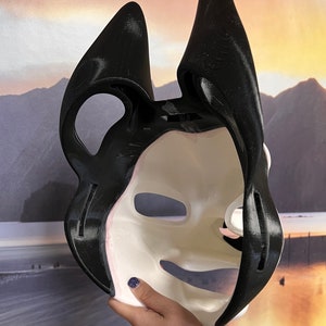 May include: A black and white mask with large, pointed ears. The mask has a smooth, off-white face with black accents and a black outline. The mask is held up, revealing the inside. The mask is likely for cosplay or costume use.