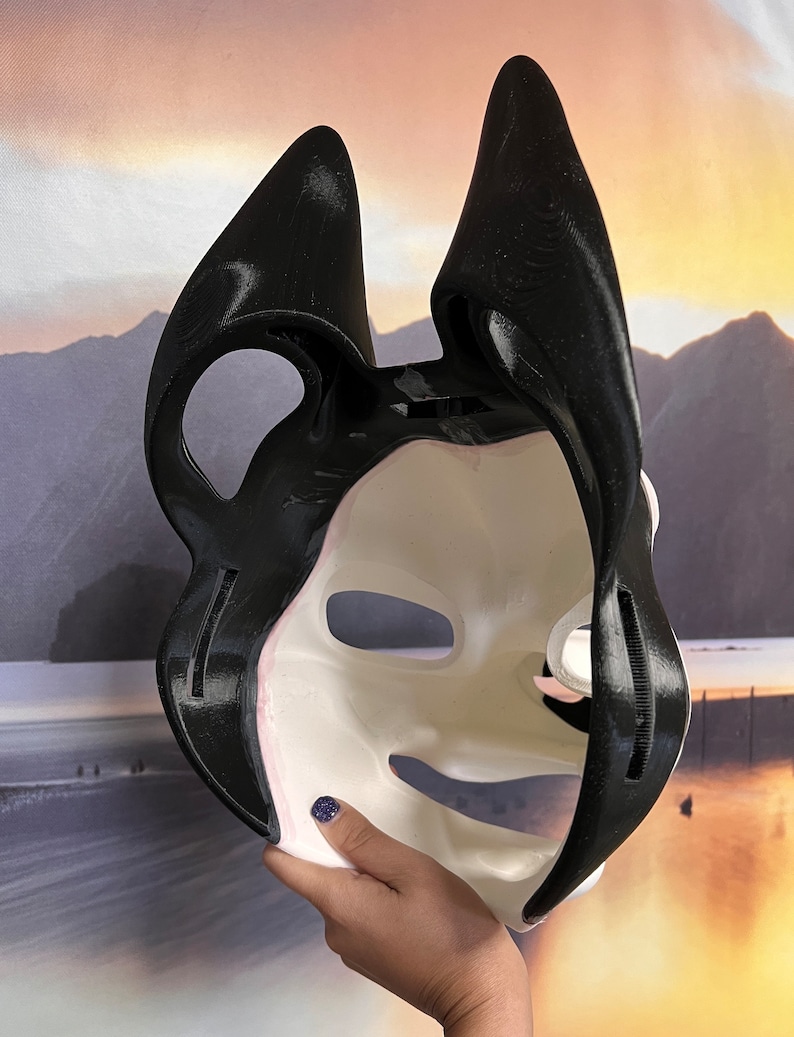 May include: A black and white mask with large, pointed ears. The mask has a smooth, off-white face with black accents and a black outline. The mask is held up, revealing the inside. The mask is likely for cosplay or costume use.