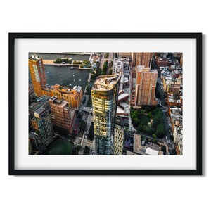 May include: An aerial photograph of a cityscape, framed in black. The image features a collection of high-rise buildings in shades of brown, gold, and grey, with a river and green areas. The photograph is a detailed view.