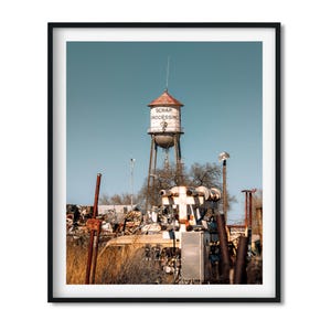 May include: A framed wall art print featuring a tall, weathered white water tower with a red roof and the text SCRAP PROCESSING printed on its side. The tower stands above a field of dry grass, industrial machinery, and a pile of scrap metal.