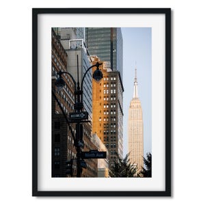 May include: Framed print of a New York City street scene. The image features the Empire State Building and other skyscrapers. A street sign reads "Ninth Ave". The buildings are a mix of brick and modern architecture.