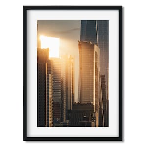 Op de afbeelding: Een foto van een skyline van een stad bij zonsondergang. De zon gaat onder achter de hoge gebouwen en werpt een warme gloed op de glazen gevels. De gebouwen zijn allemaal modern en stijlvol, met scherpe hoeken en reflecterende oppervlakken.