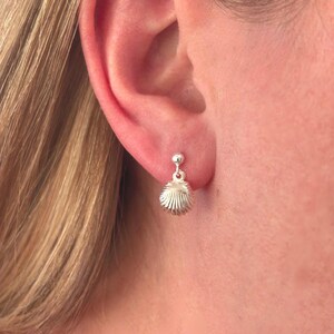 May include: A close-up of a silver earring. The earring features a small ball stud and a dangling, textured, shell-shaped charm. The earring is worn on an ear, with the ear and part of the person's hair visible.