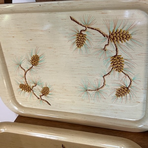 May include: Rectangular serving tray with a light beige wood grain pattern. The tray features a decorative design of brown pine cones and branches with green and blue needles. The tray has rounded edges.