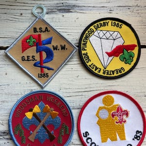 May include: Four embroidered patches featuring the Boy Scouts of America logo. The patches are circular and square in shape and feature various designs, including a diamond, crossed axes, and a scout handshake. The patches are red, blue, yellow, and white.