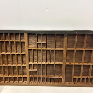 May include: A vintage wooden printer's tray with numerous small compartments. The tray is made of light-colored wood with a dark green trim. Some compartments contain letters and numbers, while others are empty. The tray is likely used for storing printing type.