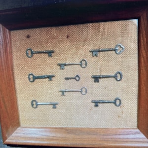 May include: A framed display of nine antique keys on a burlap background. The keys are arranged in a symmetrical pattern and are all different sizes and shapes.